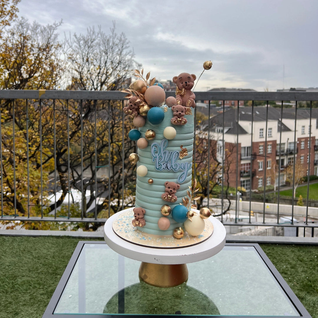 A decorated cake with the phrase 'OH BABY' displayed on top, adorned with pine cone-like decorations, presented on a glass table with a balcony view in the background.