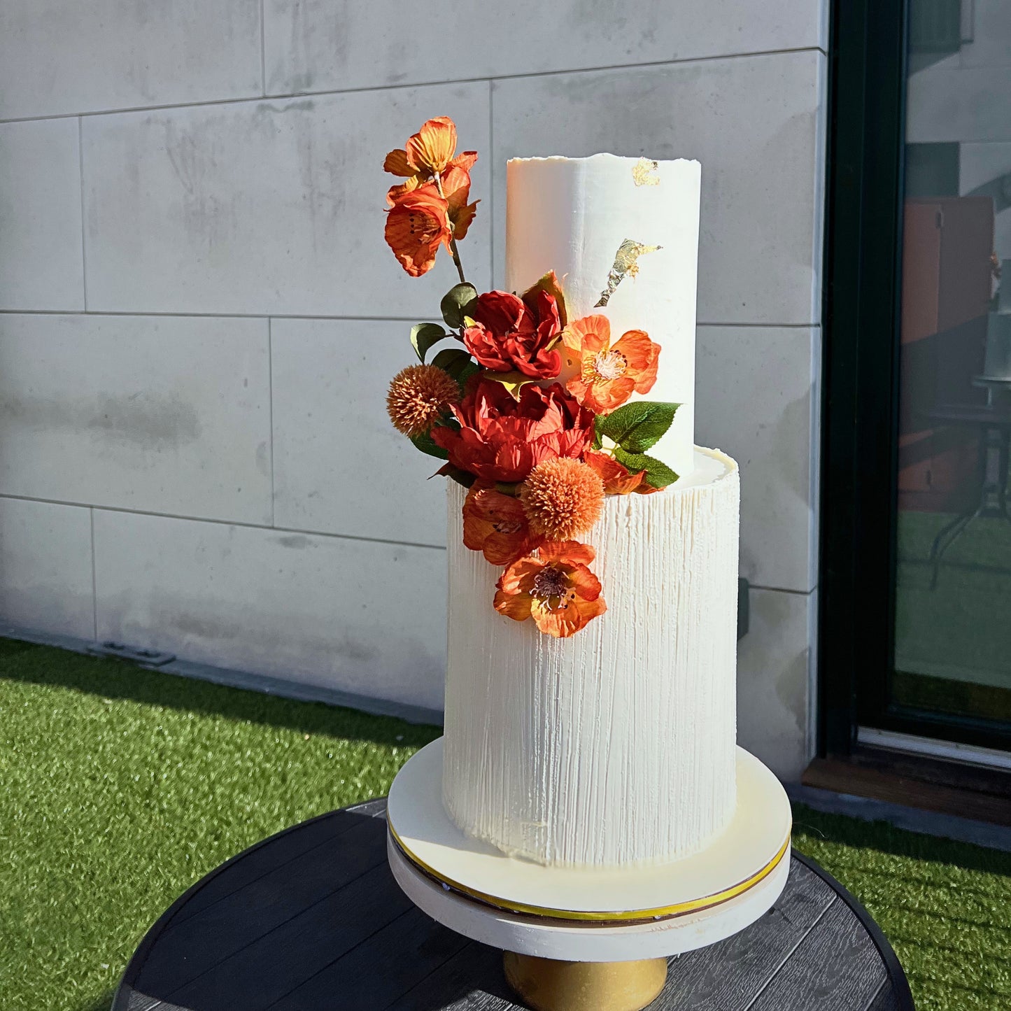 Textured ivory wedding cake with cascading burnt orange and red flowers in Dublin sunlight, CakeMeAway