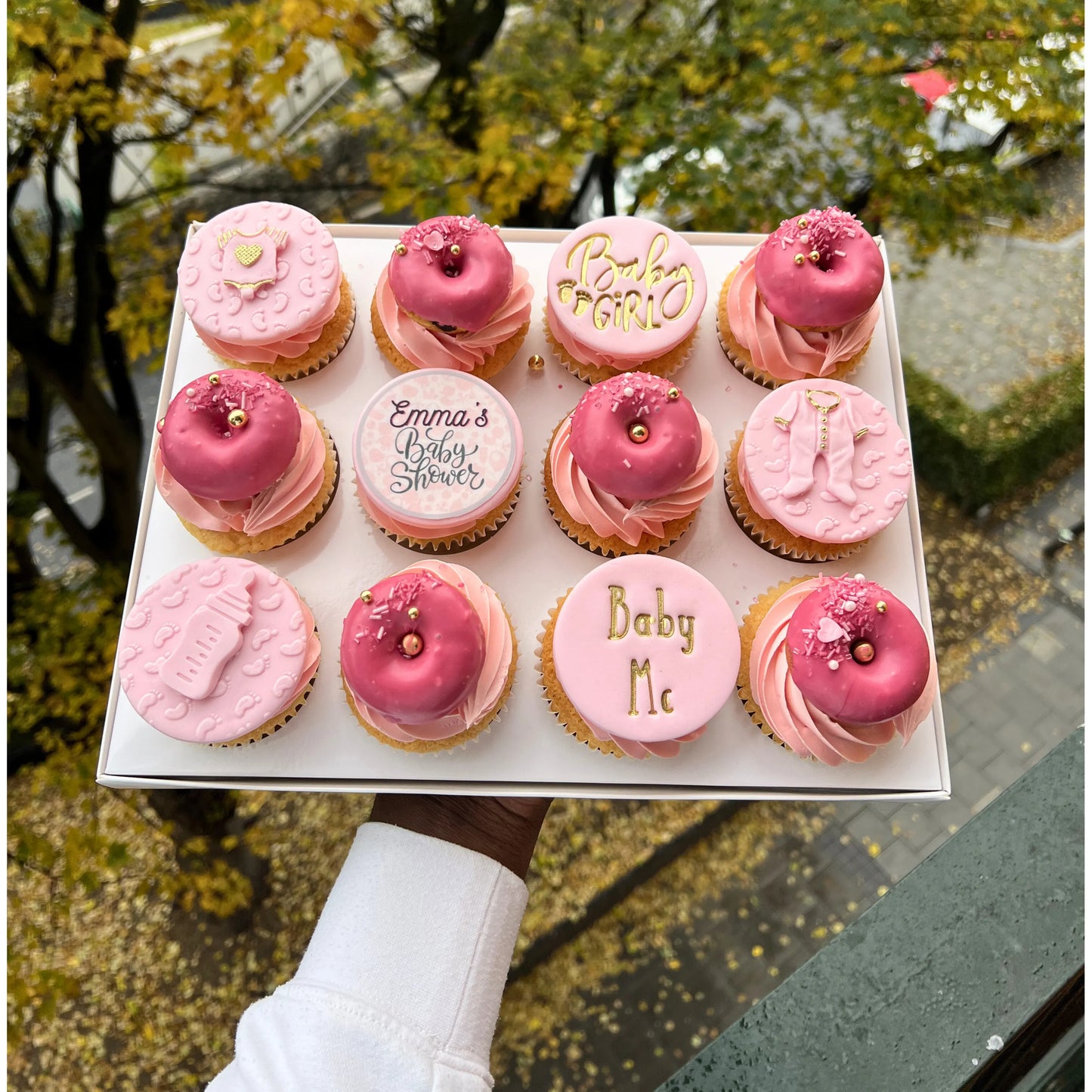 Box of pink baby shower cupcakes with baby-themed text held outdoors