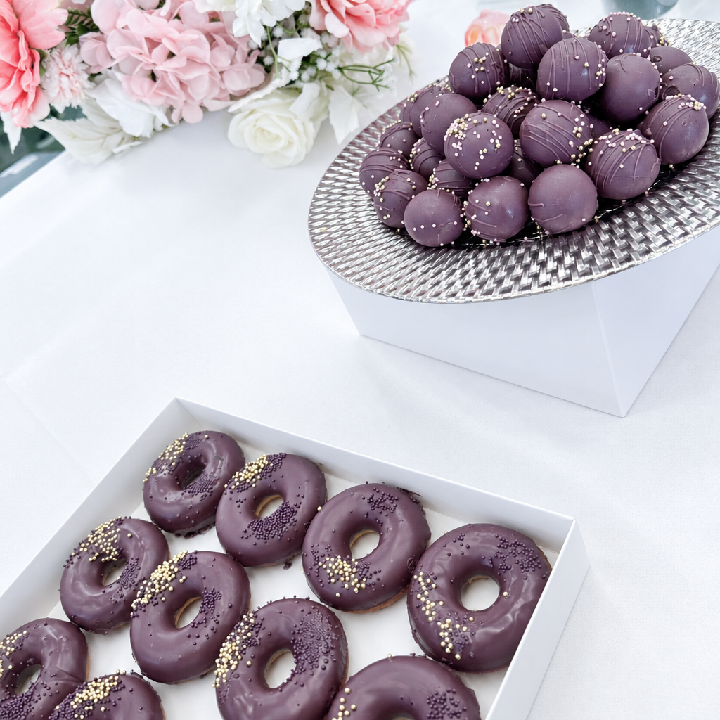 Chocolate-glazed donuts with gold sprinkles in a white box, alongside a tray of chocolate truffles on a white table with pink flowers in the background.