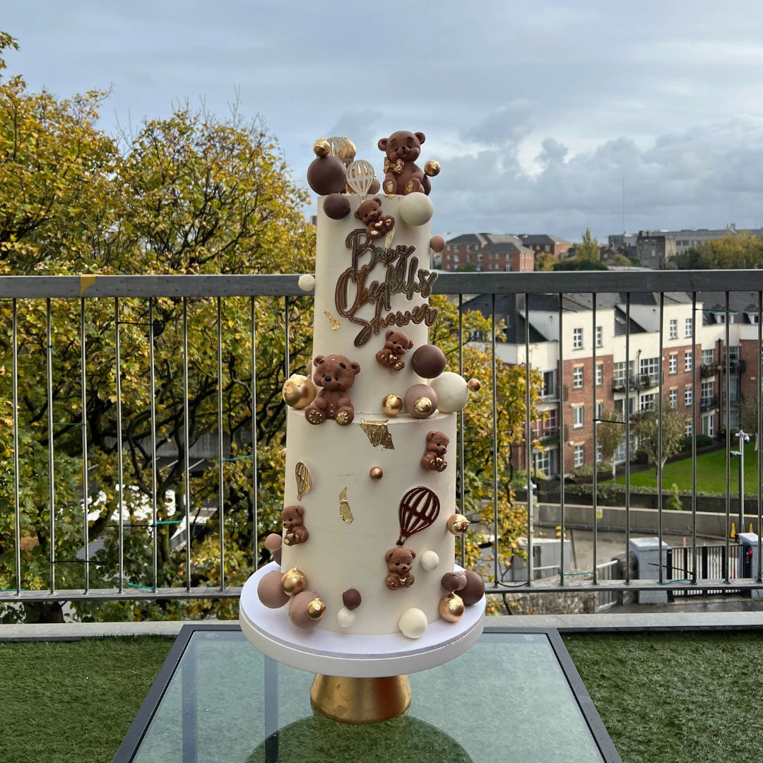 A multi-tiered baby shower cake decorated with teddy bear-themed candies and balloons, displayed on a table outdoors.