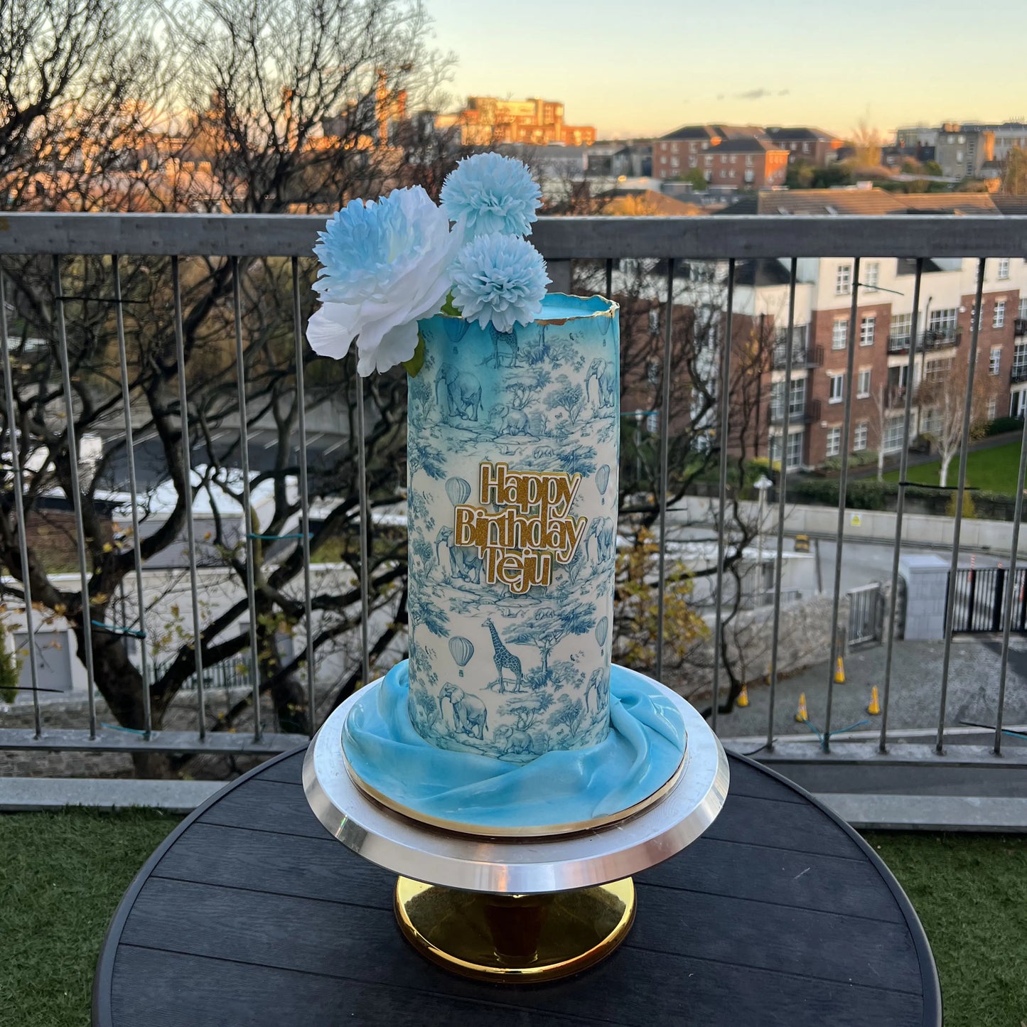 blue safari baby shower cake with baby-themed elements with floral decorations on a balcony with cityscape background