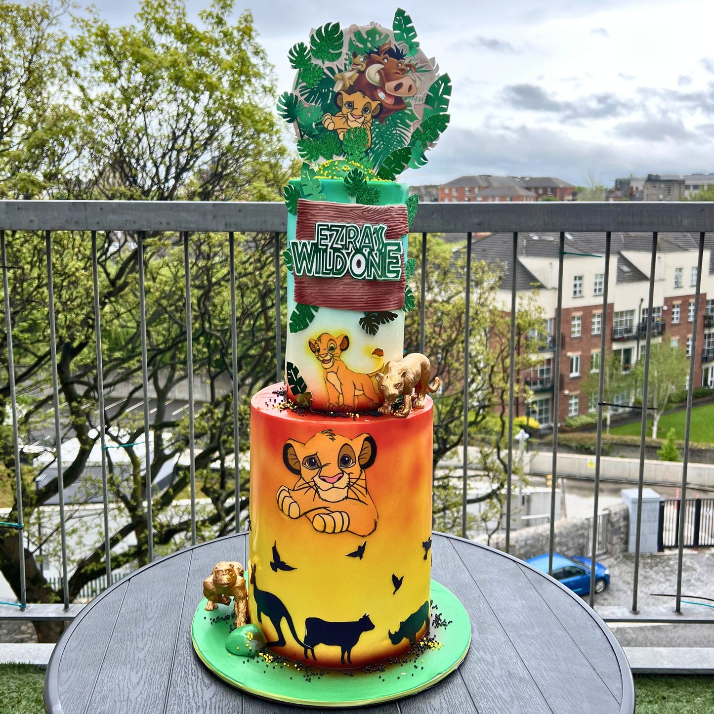 A custom two-tiered cake decorated with Simba and Nala from The Lion King, displayed on a balcony table with a city background visible through the railing.