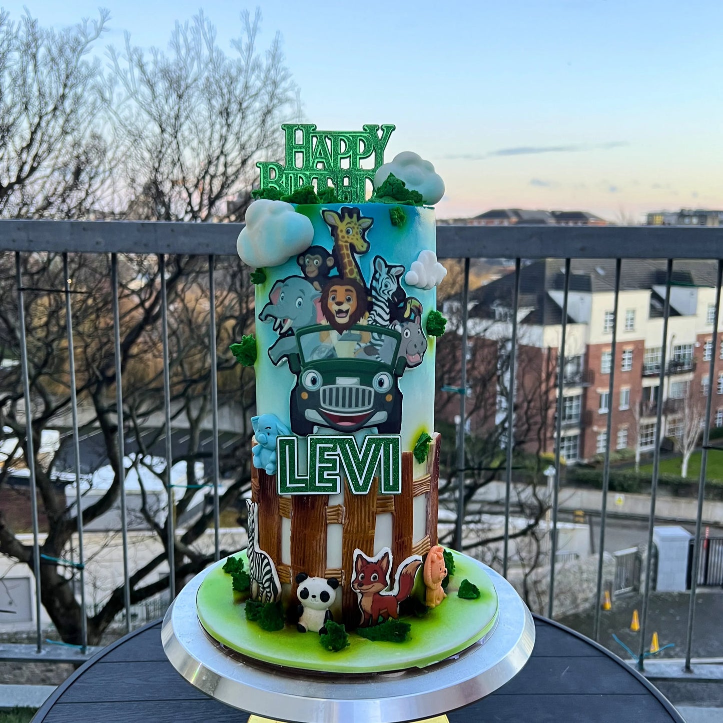 Colorful birthday cake with animal-themed decorations and 'Happy Birthday Levi' text on a balcony.