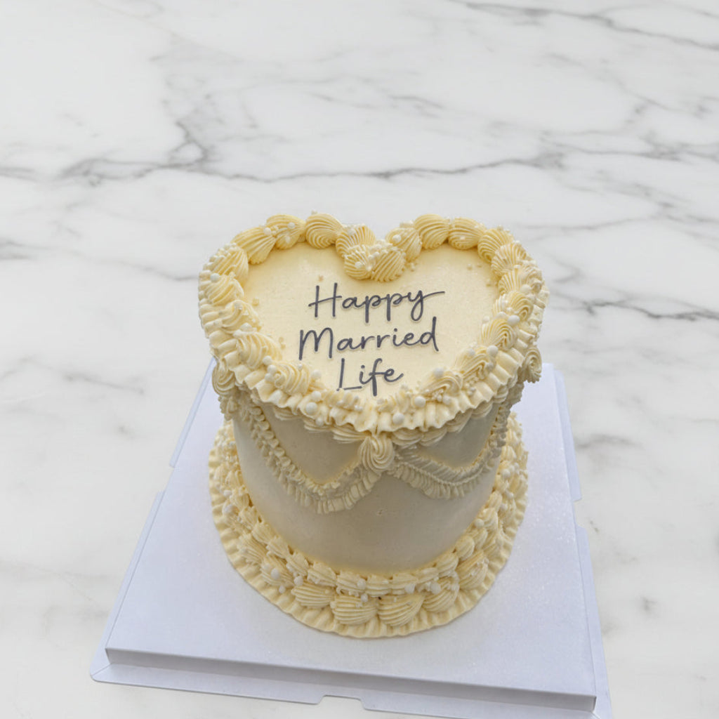 A heart-shaped frosted cake with piped borders and the words 'Happy Married Life' on top, displayed on a white cake box with an outdoor city view in the background.