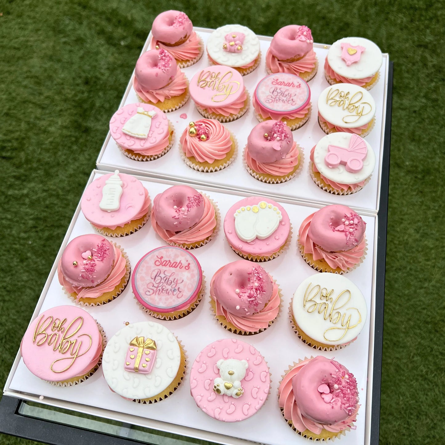 Box of pink and yellow baby shower cupcakes with pink and white icing in a box on a green grass background