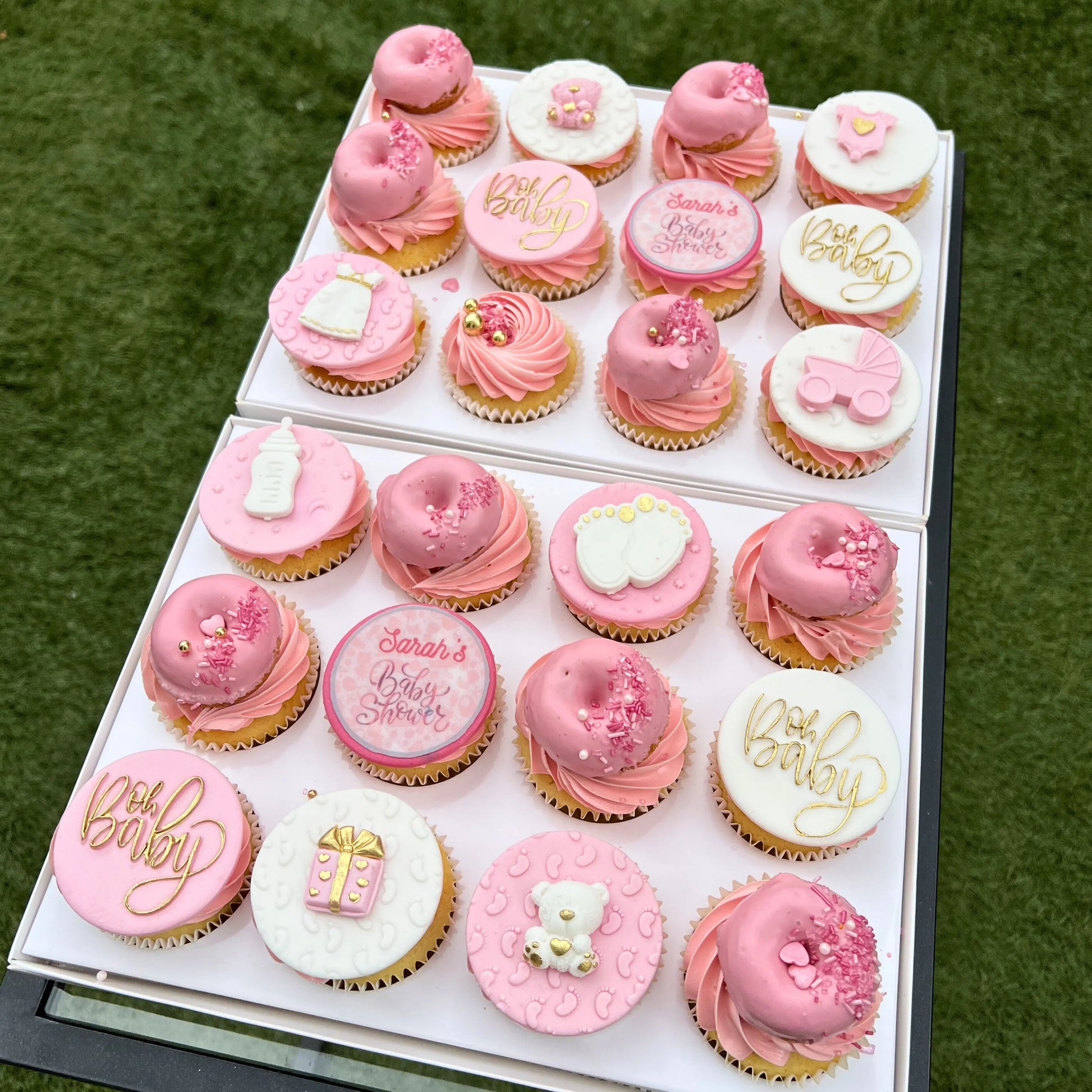 Box of pink and yellow baby shower cupcakes with pink and white icing in a box on a green grass background