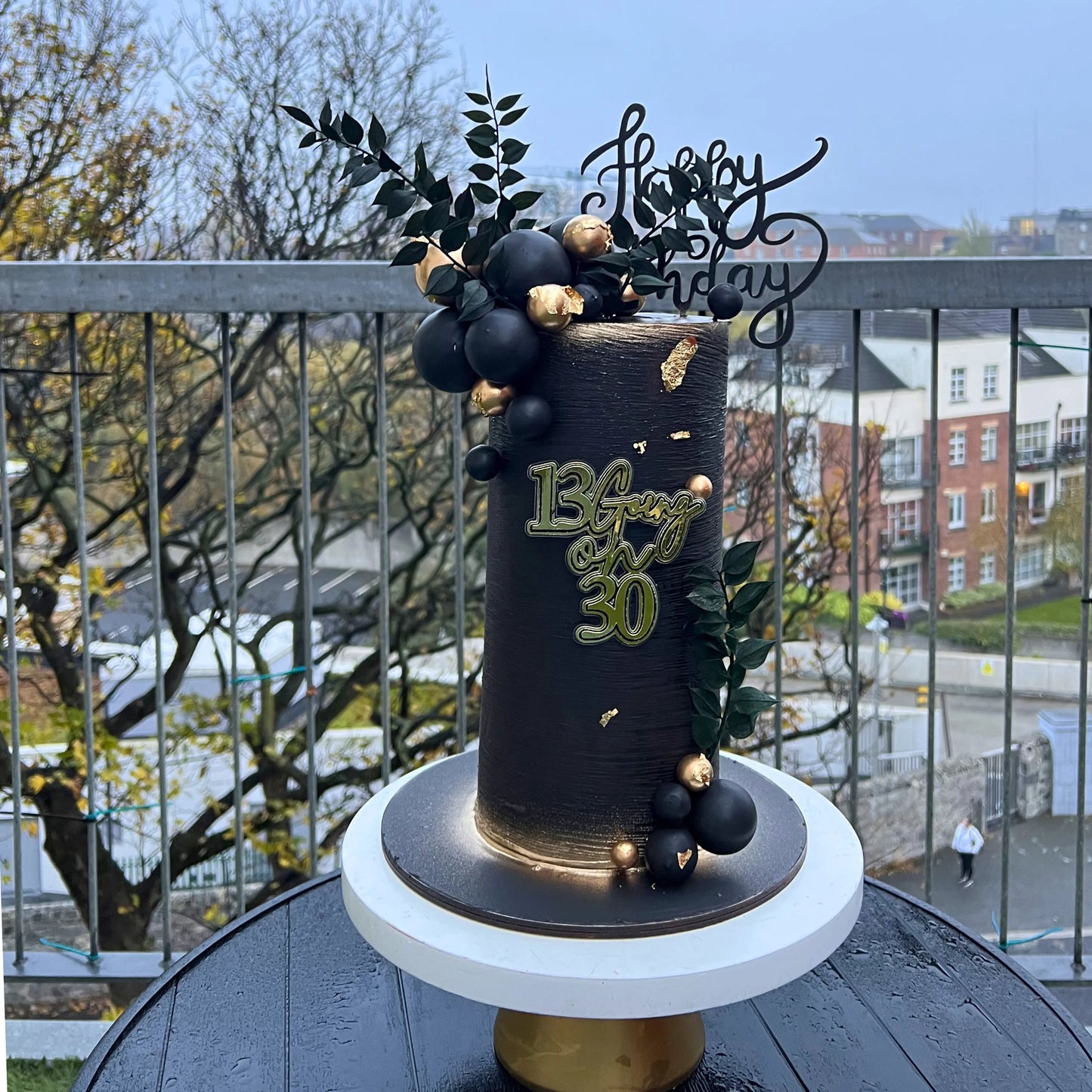 Decorative cake with gold text and black and gold decorations on a stand outdoors.