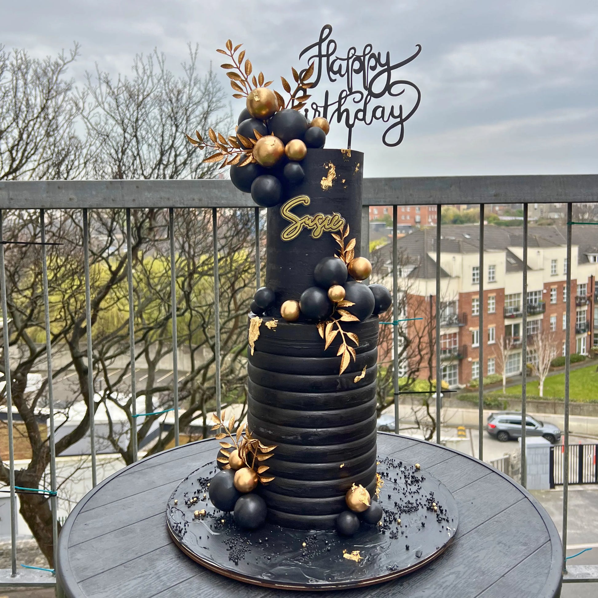 double tiered Black birthday cake with gold accents on a balcony with a cityscape background
