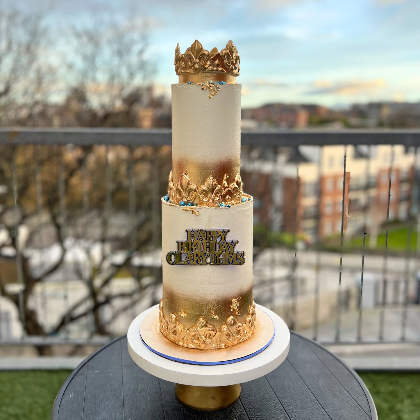 2 tiers birthday cake with gold crown and white design on a table outdoors.