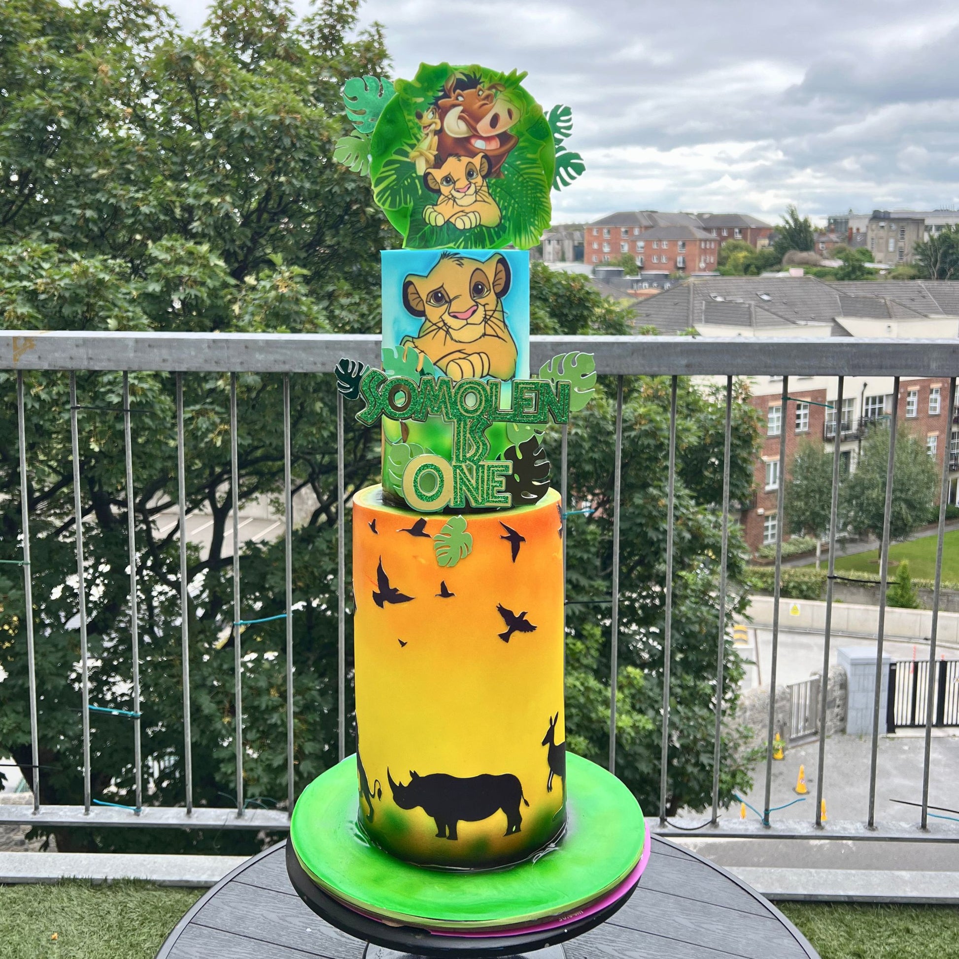 A custom two-tiered cake decorated with a vibrant yellow-to-orange gradient, black animal silhouettes, and a green base, topped with a green jungle-themed lion cub topper and a 'SONYOLEN TO BE ONE' sign. The cake is displayed on a balcony with trees and buildings in the background.
