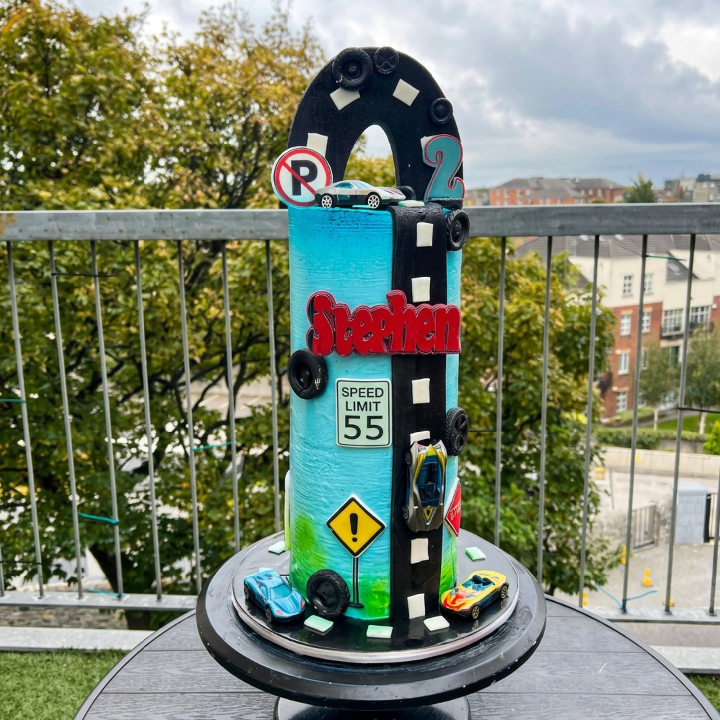 Tall turquoise race car birthday cake with fondant racetrack, speed limit sign and toy cars, handmade in Dublin by CakeMeAway