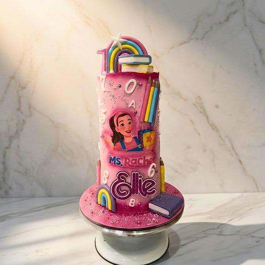 Tall pink 1st birthday cake with pastel rainbow, fondant letters, numbers, coloured pencils and books, CakeMeAway Dublin