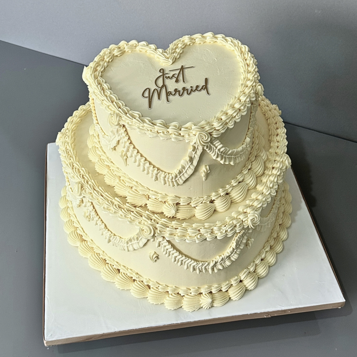 A 2-tier heart-shaped wedding cake with cream-colored frosting, piped decorative borders, and 'Just Married' written on the top tier, displayed on a white square cake board against a gray background.