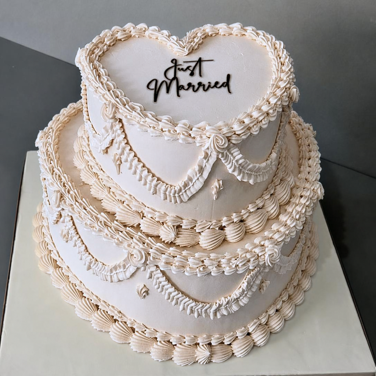 A 2 tiered white wedding cake decorated with piped cream frosting borders and scrollwork, topped with a heart-shaped section that reads 'Just Married' in black script.