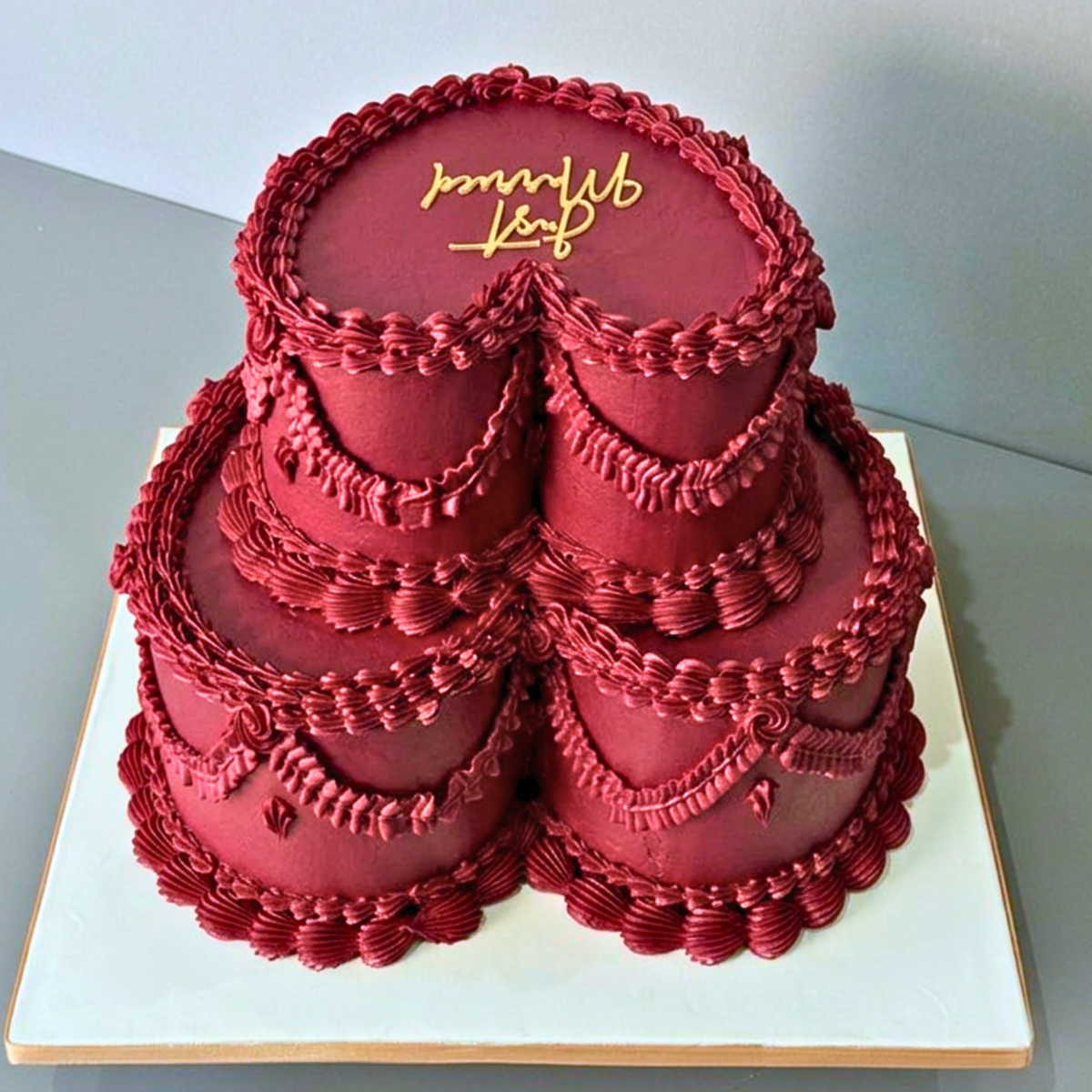 A 2-tier heart-shaped wedding cake decorated in deep red frosting with piped borders and gold script reading 'Jonathan & Beth' on the top tier, displayed on a white square board against a gray background.