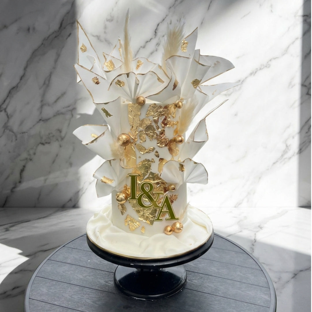 A custom white and gold tiered cake with decorative gold leaf, feathers, and a monogram topper reading 'I&A', displayed on a black cake stand against a marble-patterned background.