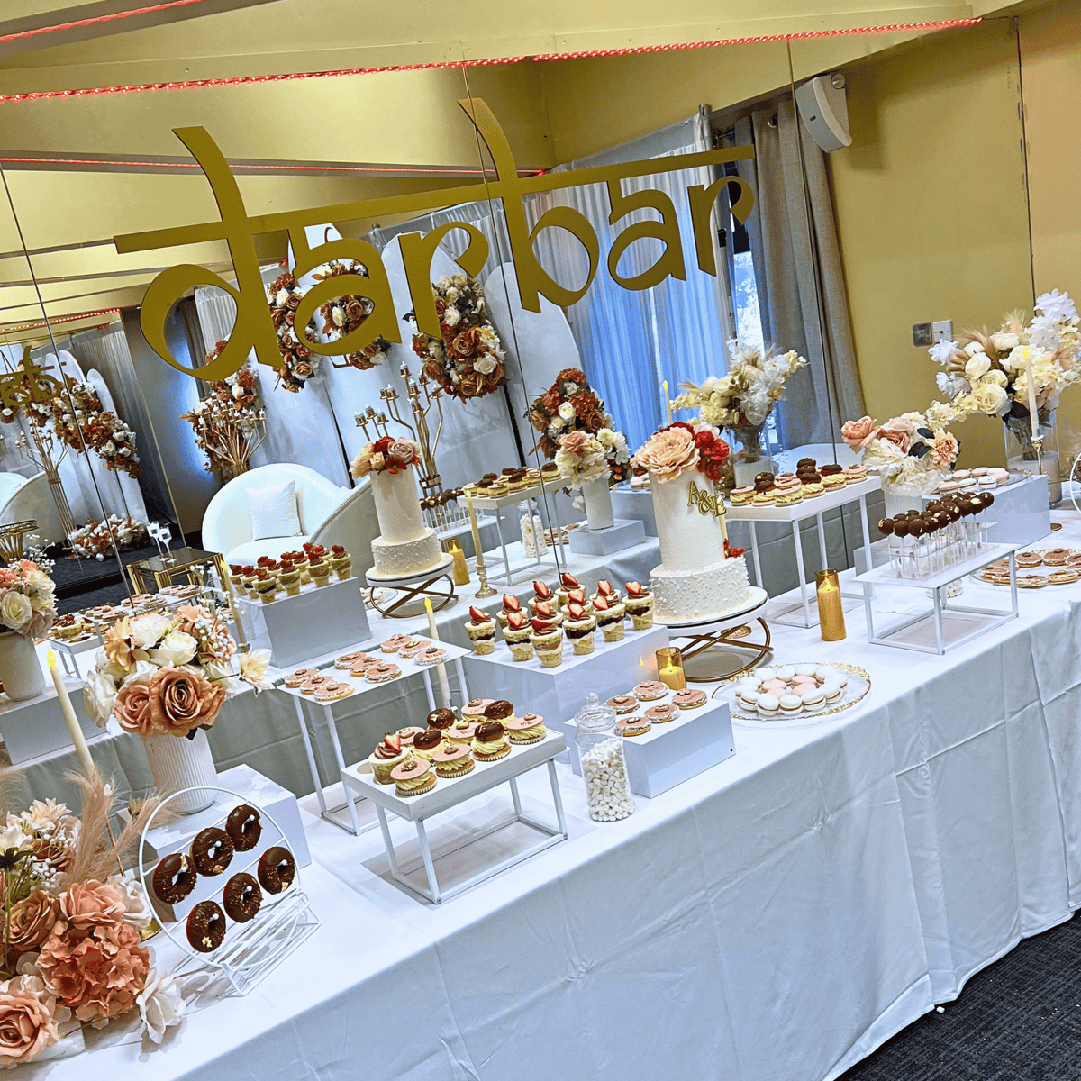 A decorated dessert table at a celebration or event, featuring cupcakes, pastries, flowers, and a large gold hanging sign that reads 'darbar' against a mirrored backdrop.