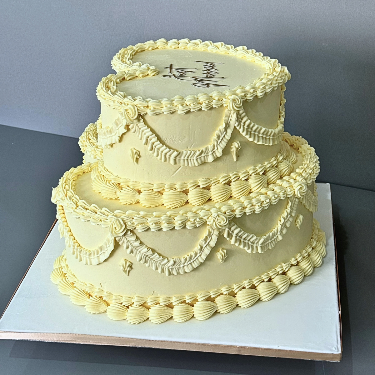 A two-tiered round cake decorated with pale yellow frosting and piped decorative borders, shown on a white square board against a gray background.