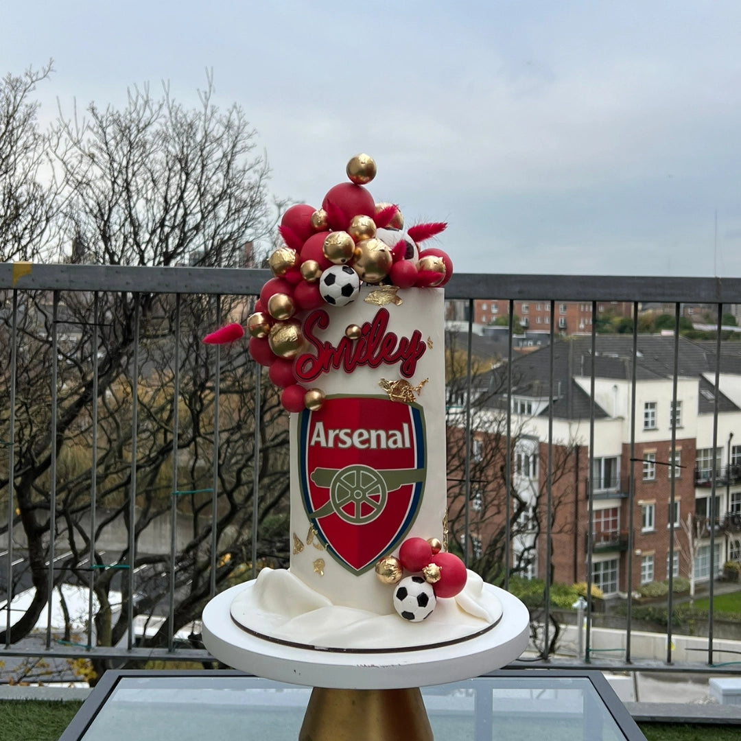 A decorated cake with Arsenal Football Club colors and themed decorations, including balloons and a topper.