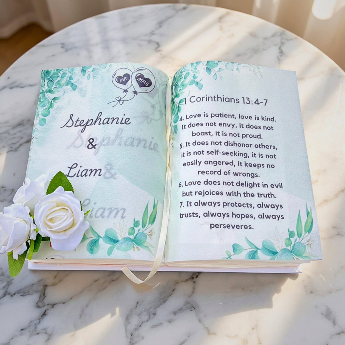 An open personalized Bible bookmark cake on a white marble surface, featuring floral watercolor designs, the names Stephanie & Liam, and a ribbon bookmark. The left page is decorated with white roses, and the right page displays a Bible passage from 1 Corinthians 13:4-7.