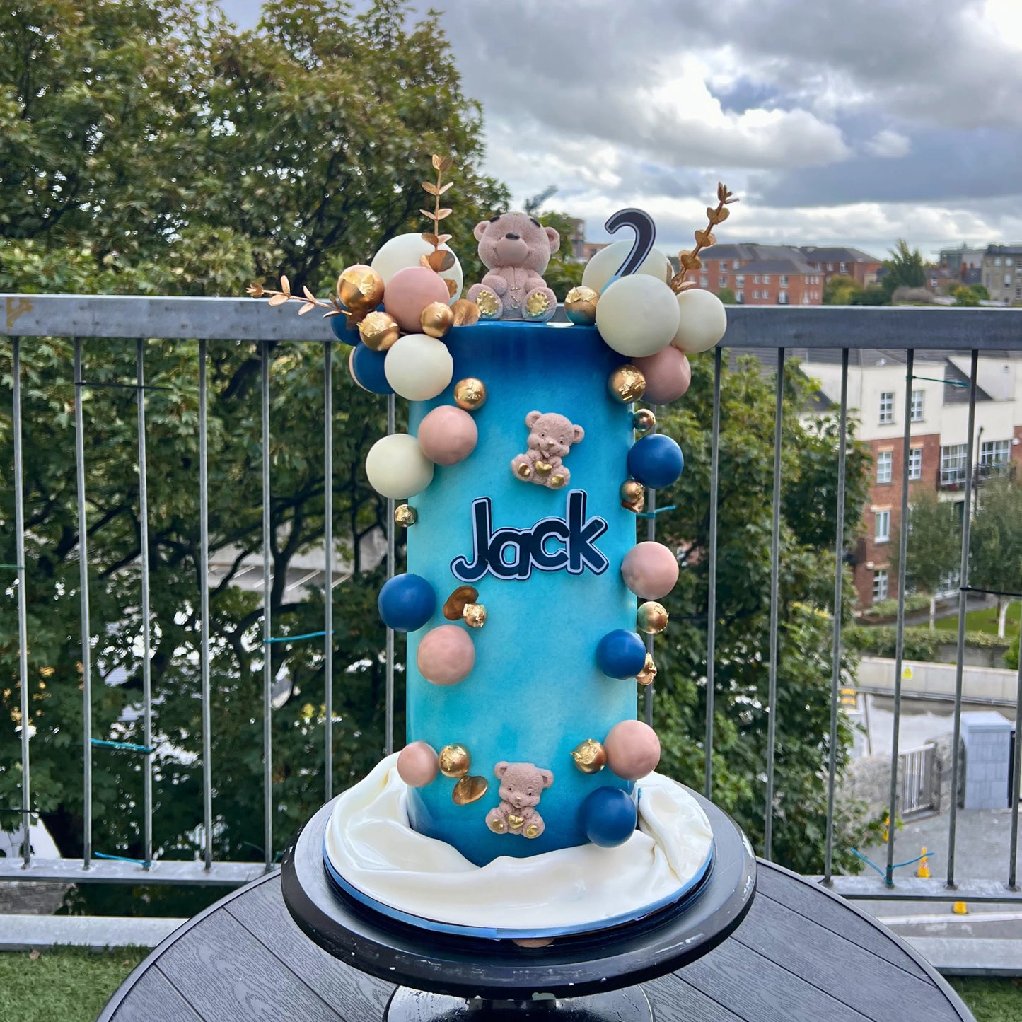 Blue cake with teddy bears and balloons on a balcony with trees and buildings in the background