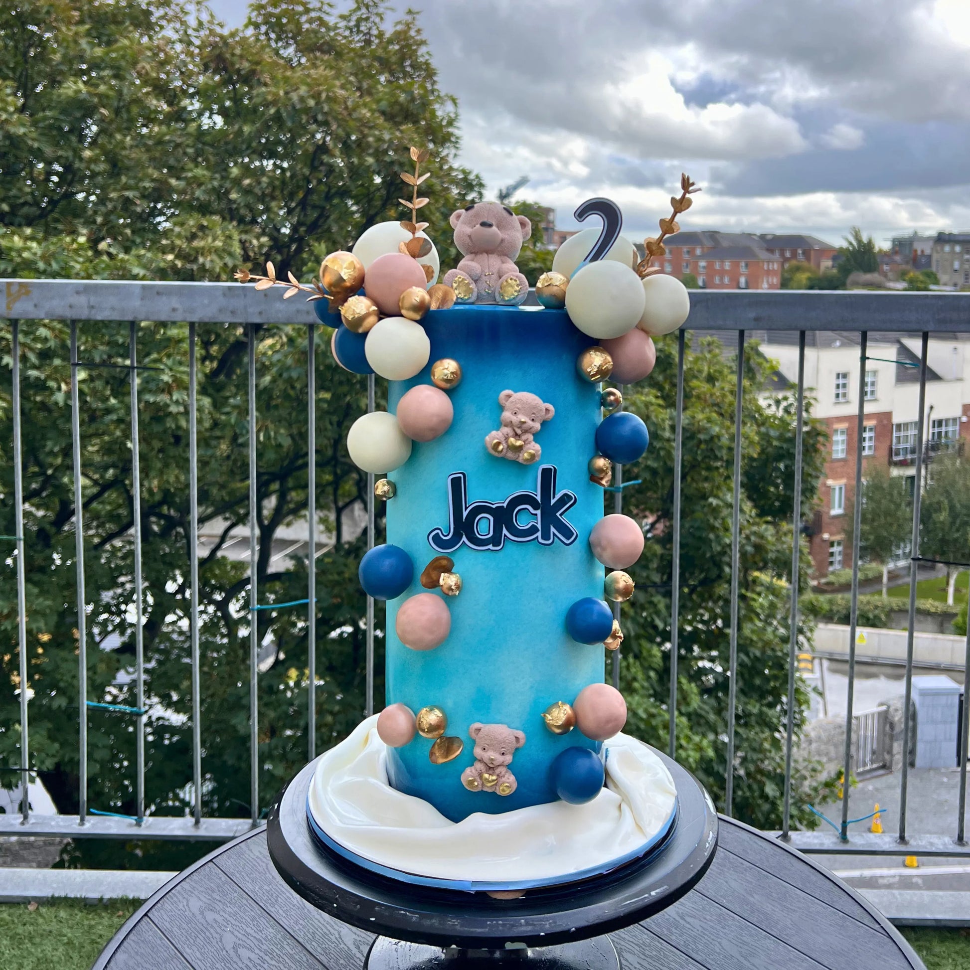 Blue cake with teddy bears and balloons on a balcony with trees and buildings in the background