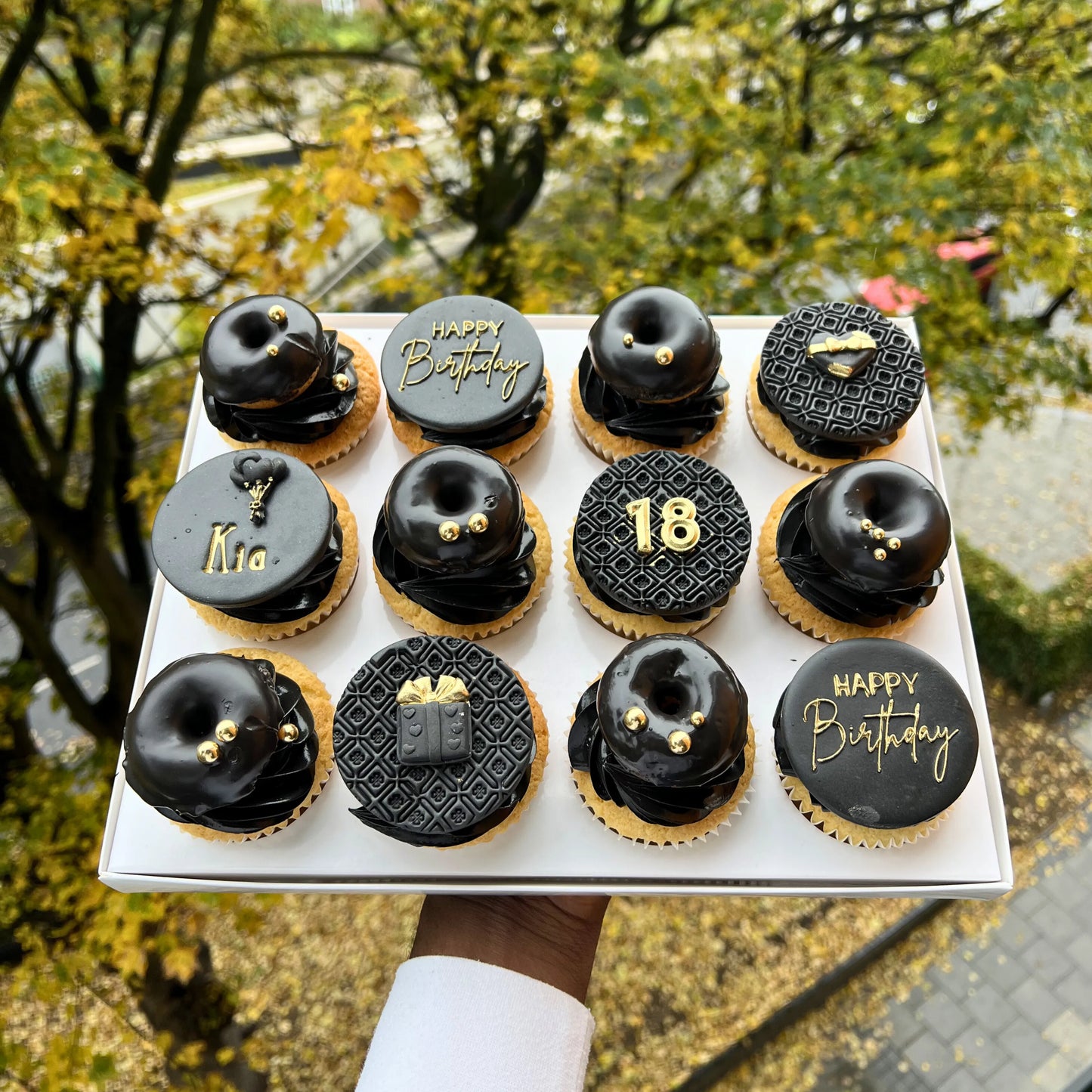 Box of black and gold birthday-themed cupcakes outdoors