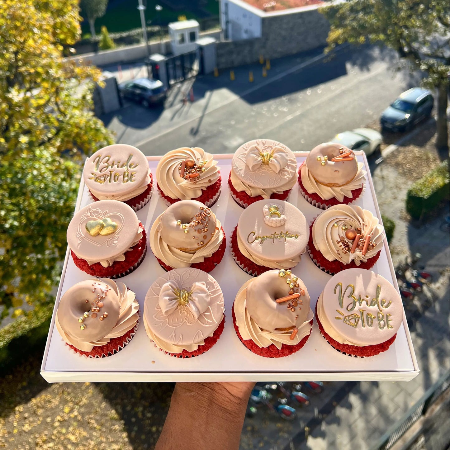 Box of cupcakes with 'Bride to be' and other decorative elements held outdoors.