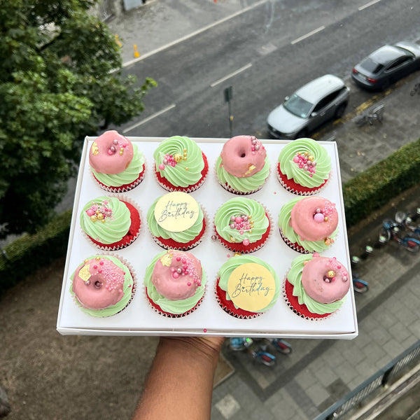 Box of cupcakes with pink and green frosting, one displaying 'Happy Birthday' message, held outdoors.