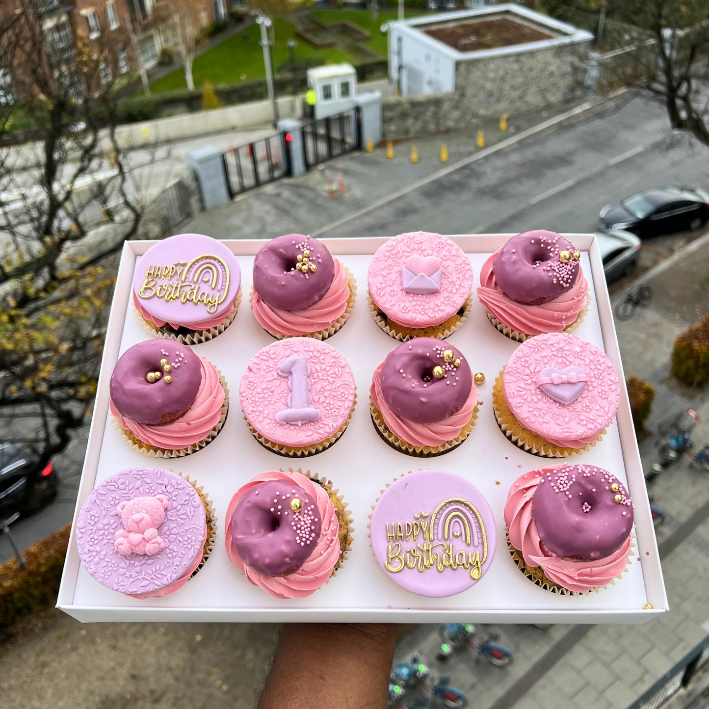 Box of pink and purple cupcakes with birthday decorations held up against an urban background.