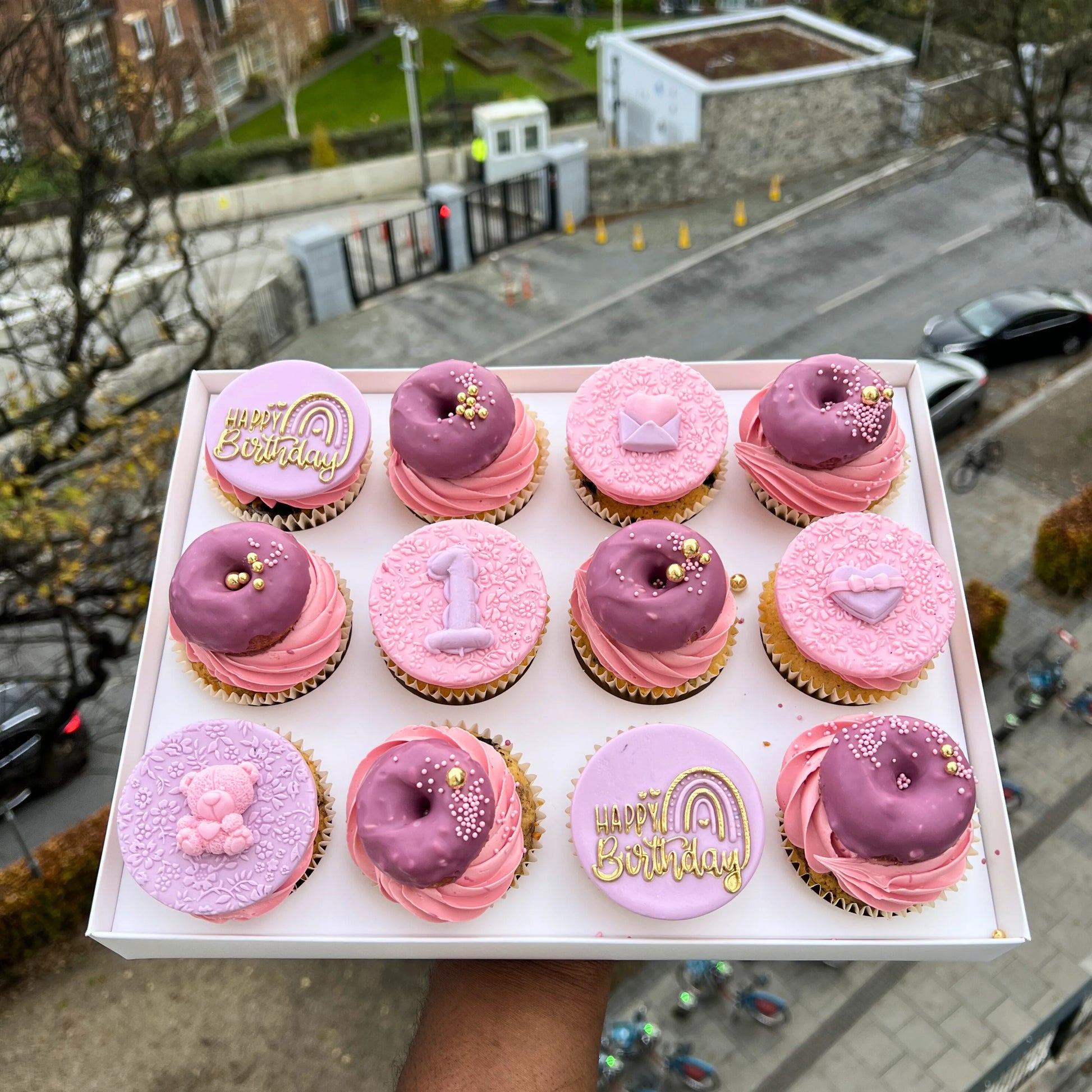 Box of pink and purple cupcakes with birthday decorations held up against an urban background.