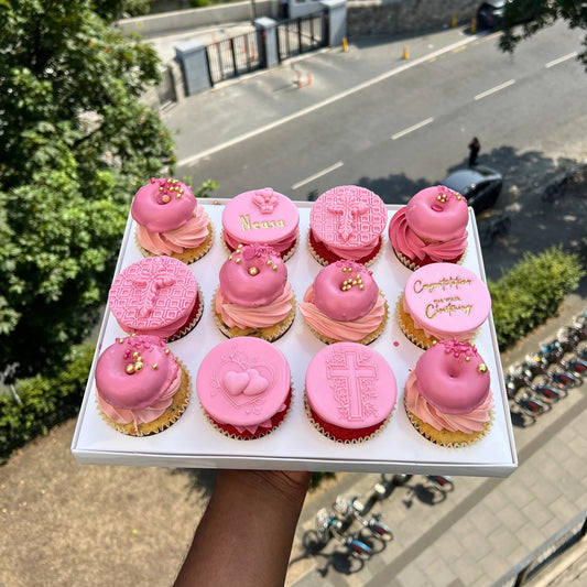 Box of pink communion cupcakes with gold accents held above a street scene