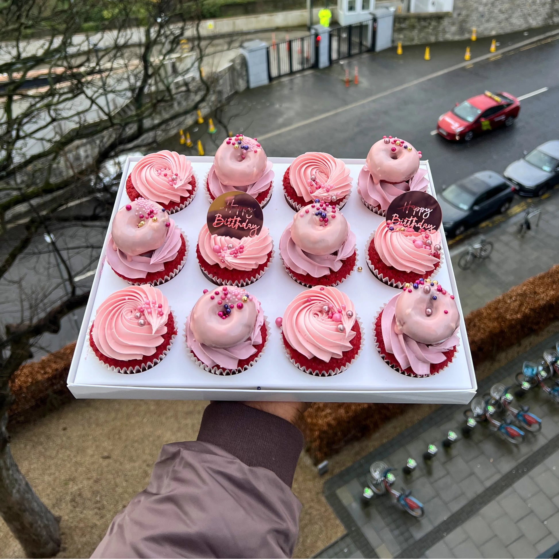 Box of pink cupcakes with red frosting held above a street scene