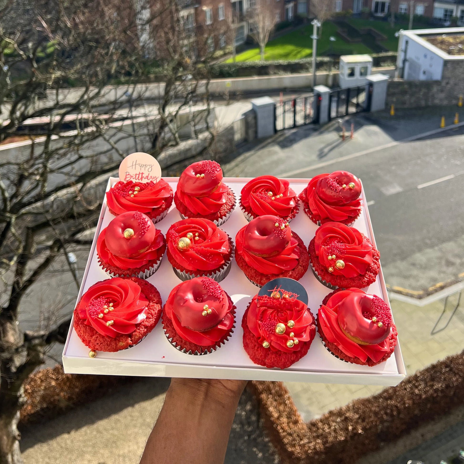 Box of red cupcakes with gold decorations held outdoors.