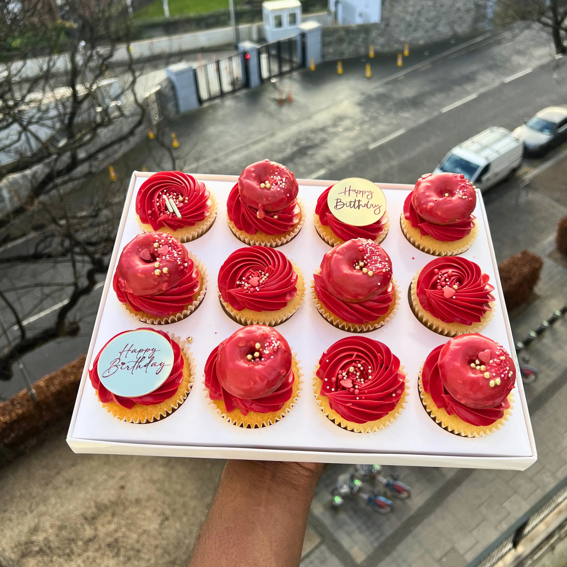 Box of red frosted cupcakes with gold accents held outdoors.
