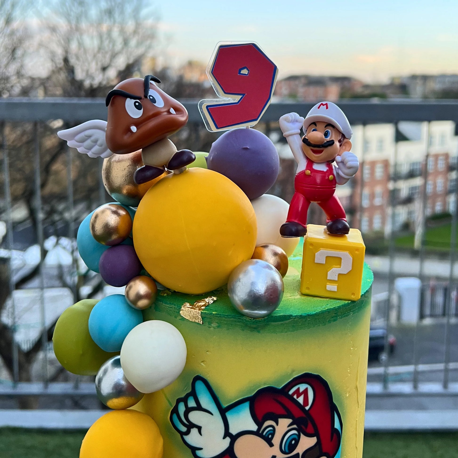 Colorful birthday cake with Mario and Goomba figures and a number 9 candle, outdoors.