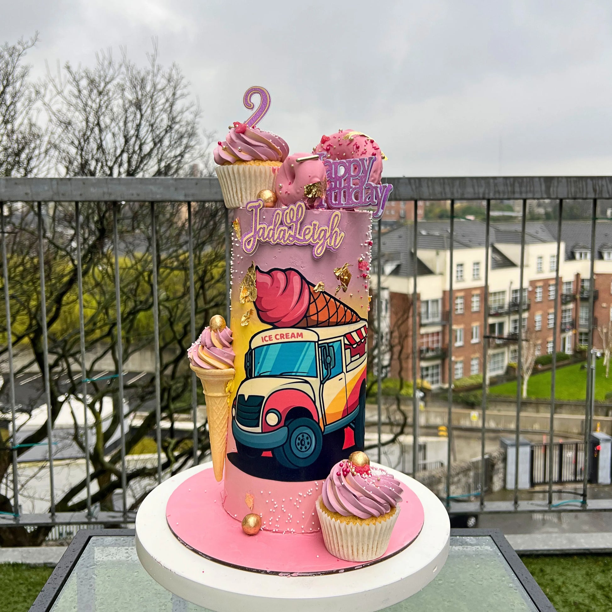Colorful birthday cake with ice cream truck design on a rooftop with buildings in the background