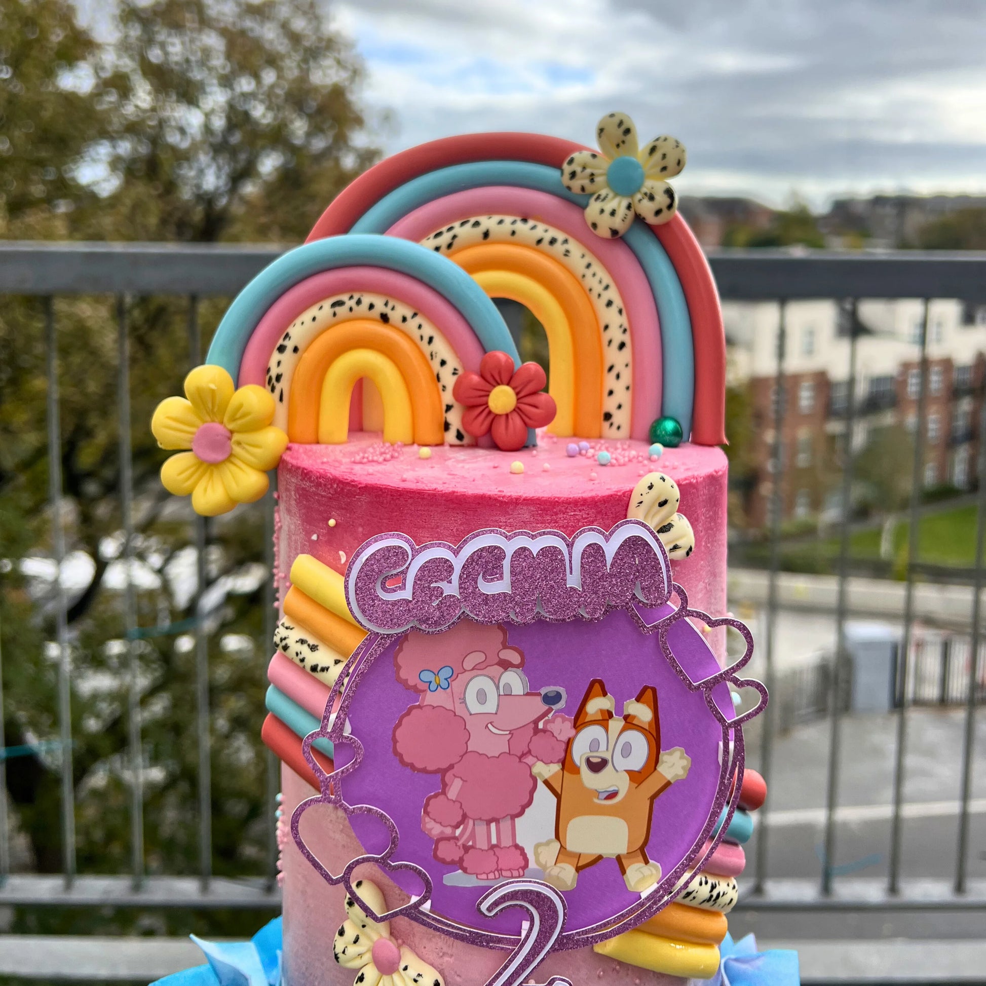 Colorful bluey birthday cake with cartoon characters and decorations on a balcony.