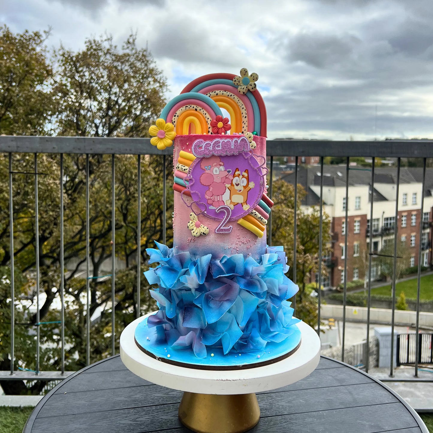 Colorful bluey birthday cake with rainbow and flowers on a stand outdoors.