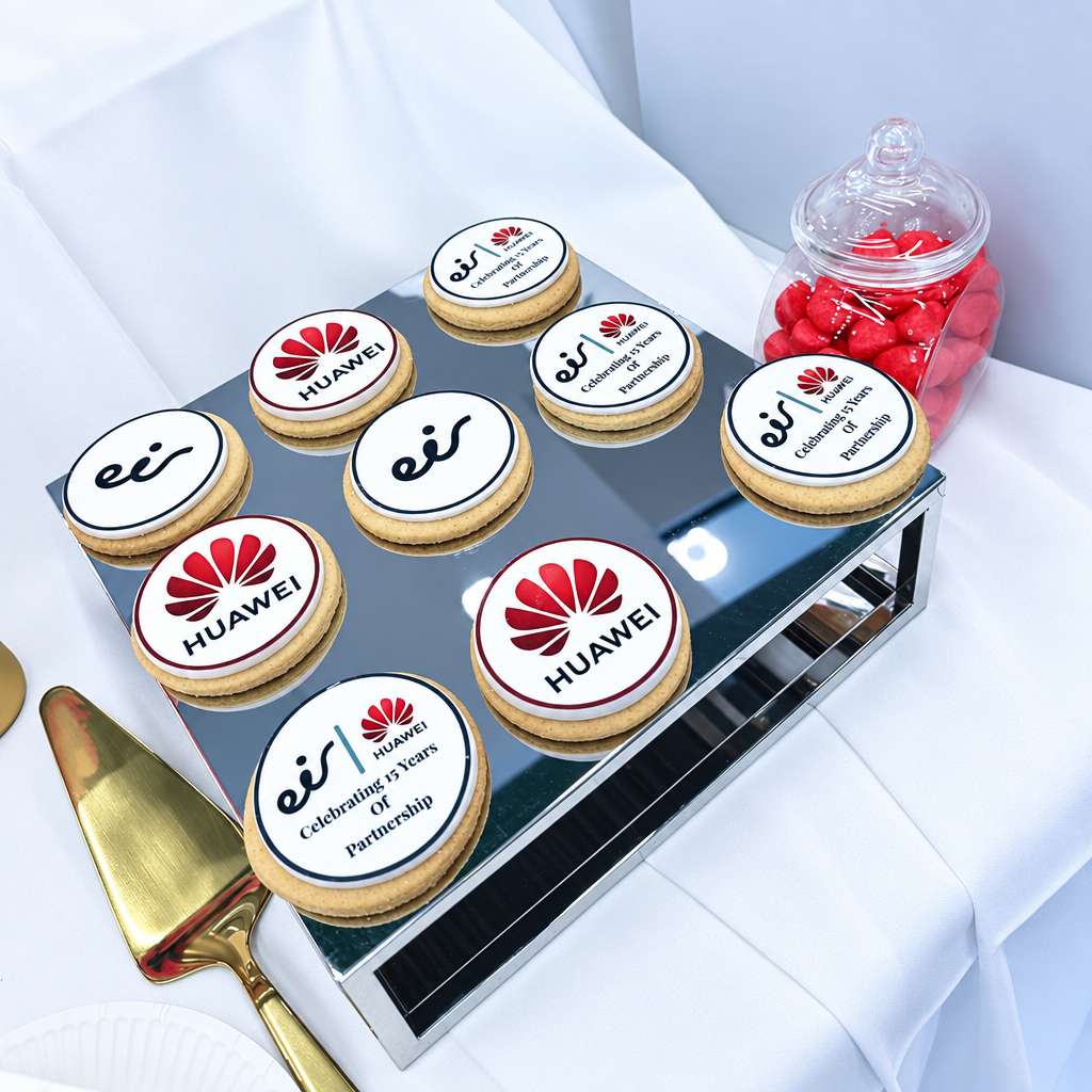 A mirrored tray holding eight round Huawei-branded cookies or tokens, each topped with a white label featuring the red Huawei flower logo and text. The tray sits on white fabric with a gold serving spatula nearby and a glass jar of red candies at the upper right.