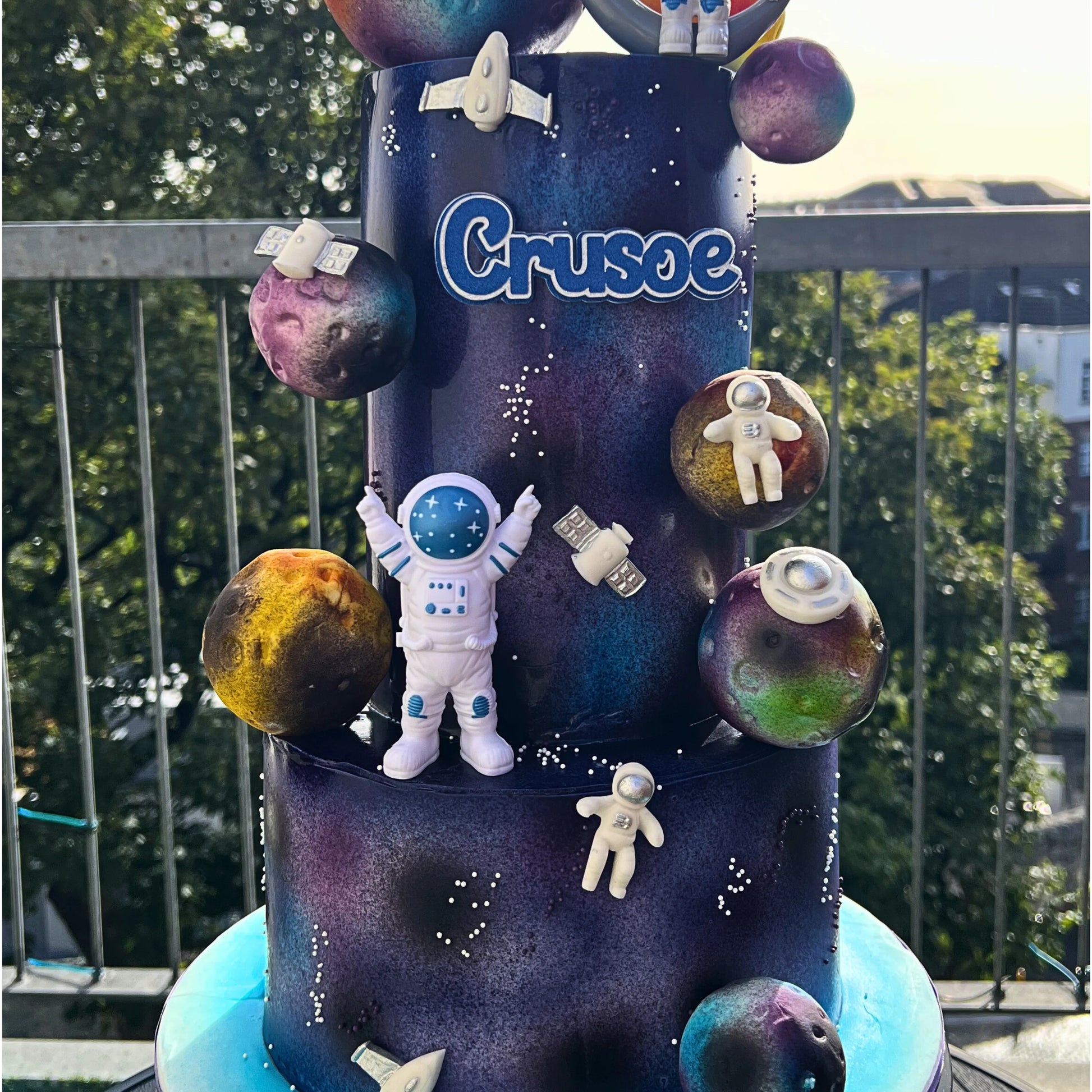 Toy tower cake with space-themed figures and 'Crusoe' branding against a blurred outdoor background