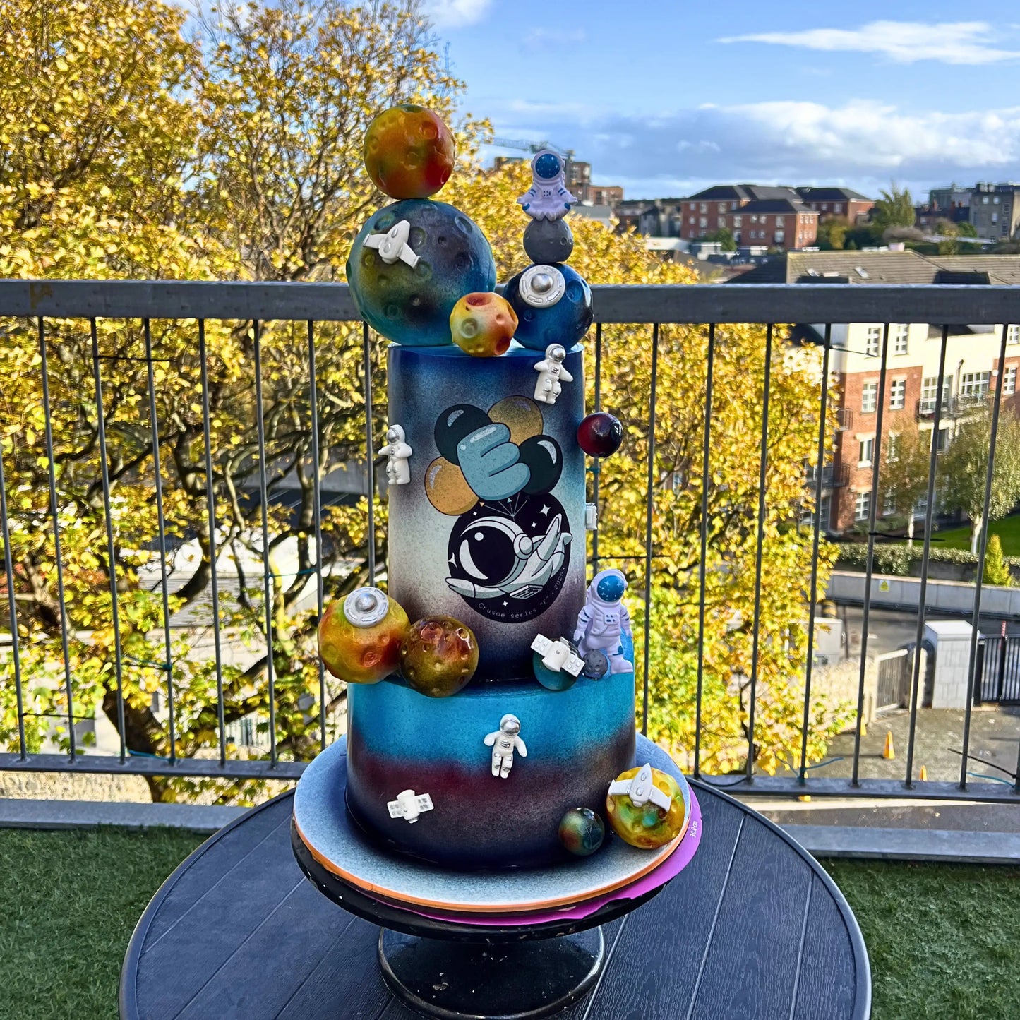 Colorful space-themed cake on a table with a cityscape background