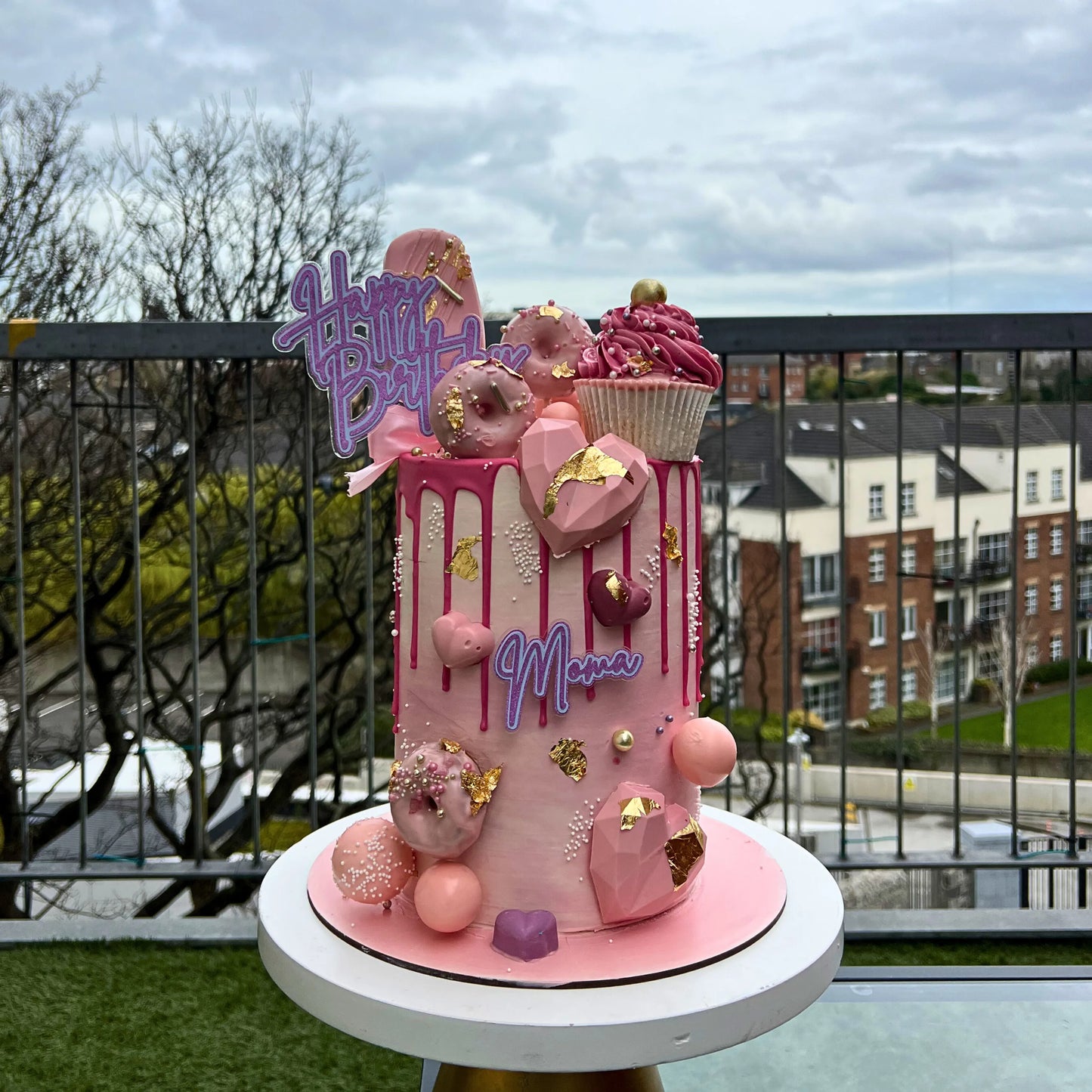 Custom birthday cake with pink icing, donuts, and 'Happy Birthday' sign on a rooftop with cityscape background.