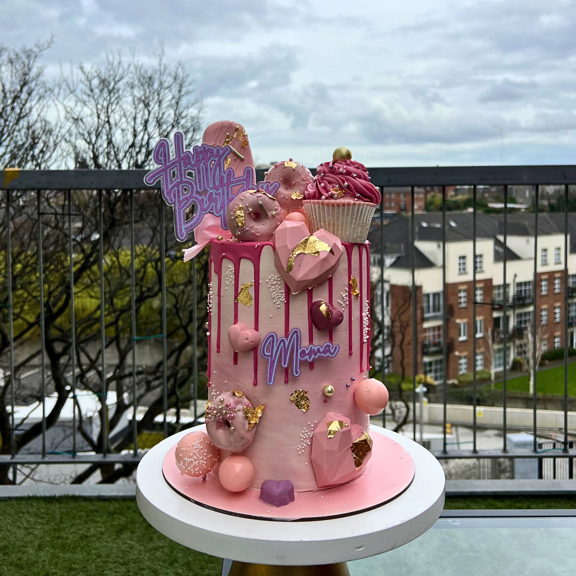Custom birthday cake with pink icing, donuts, and 'Happy Birthday' sign on a rooftop with cityscape background.