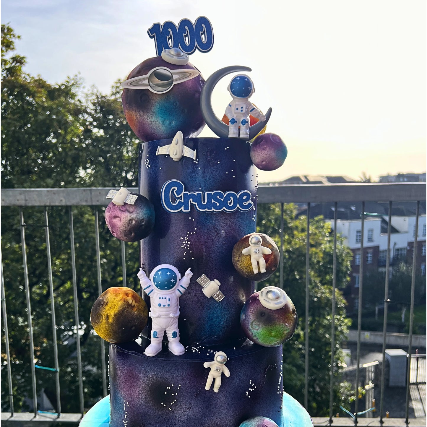 Fondant space-themed cake with space figurines, and planets against a blurred outdoor background.