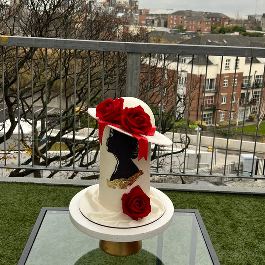 A decorated fondant cake designed to resemble a woman's head wearing a red hat, with a gold band and a red rose on top, set against an outdoor backdrop.