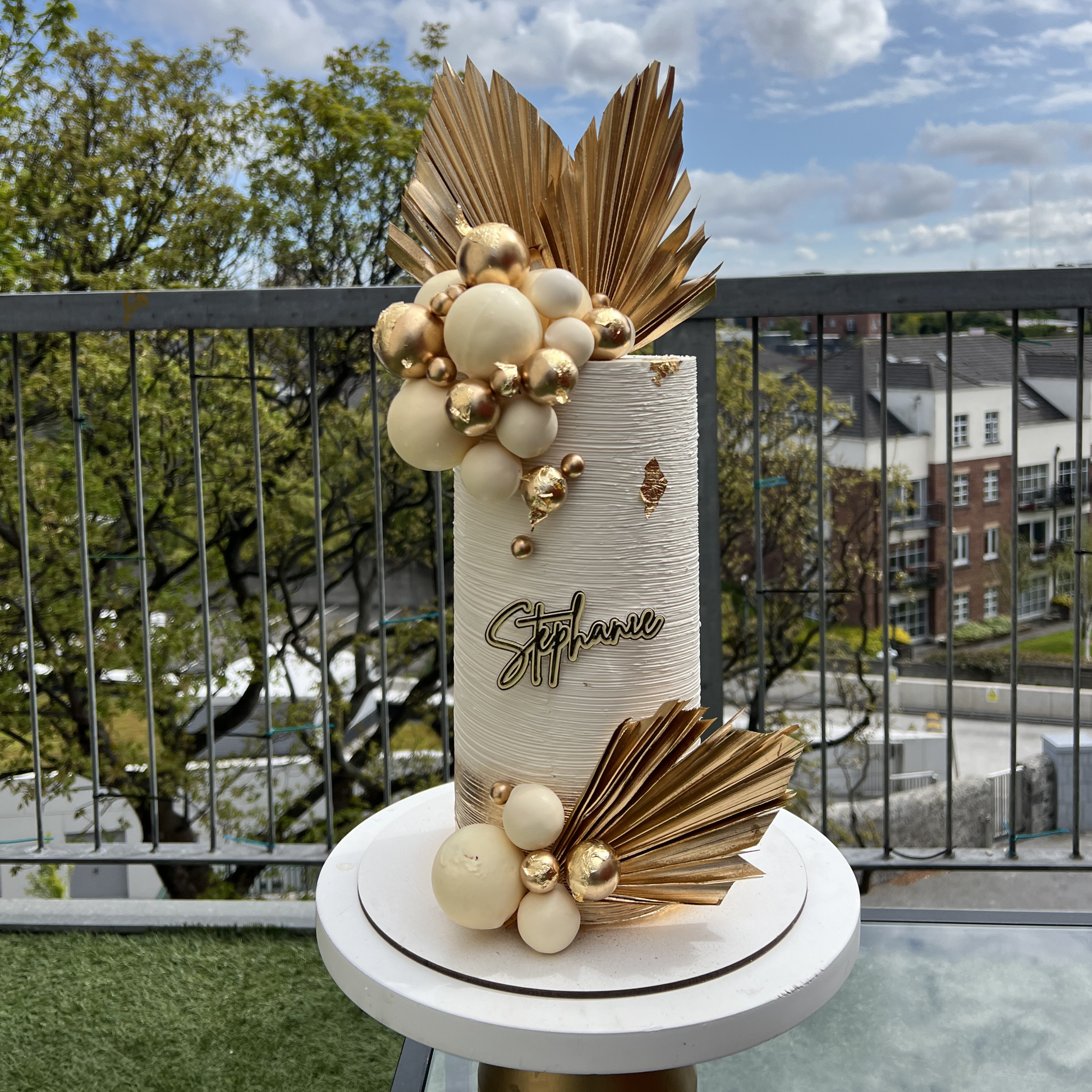 Decorative cake with gold and white elements on a stand outdoors.