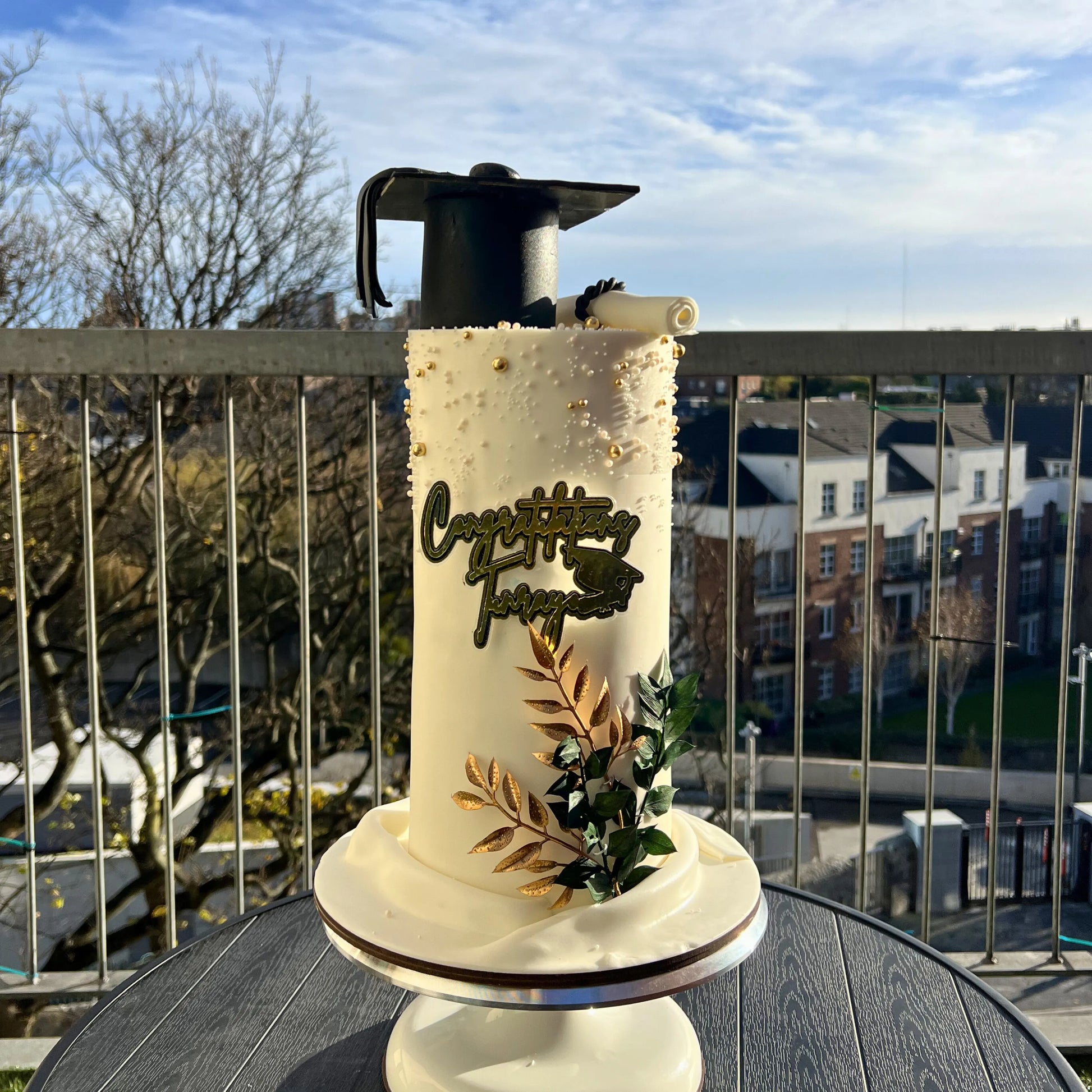 Decorative graduation cake with 'Congratulations' text on a balcony with a view of buildings.