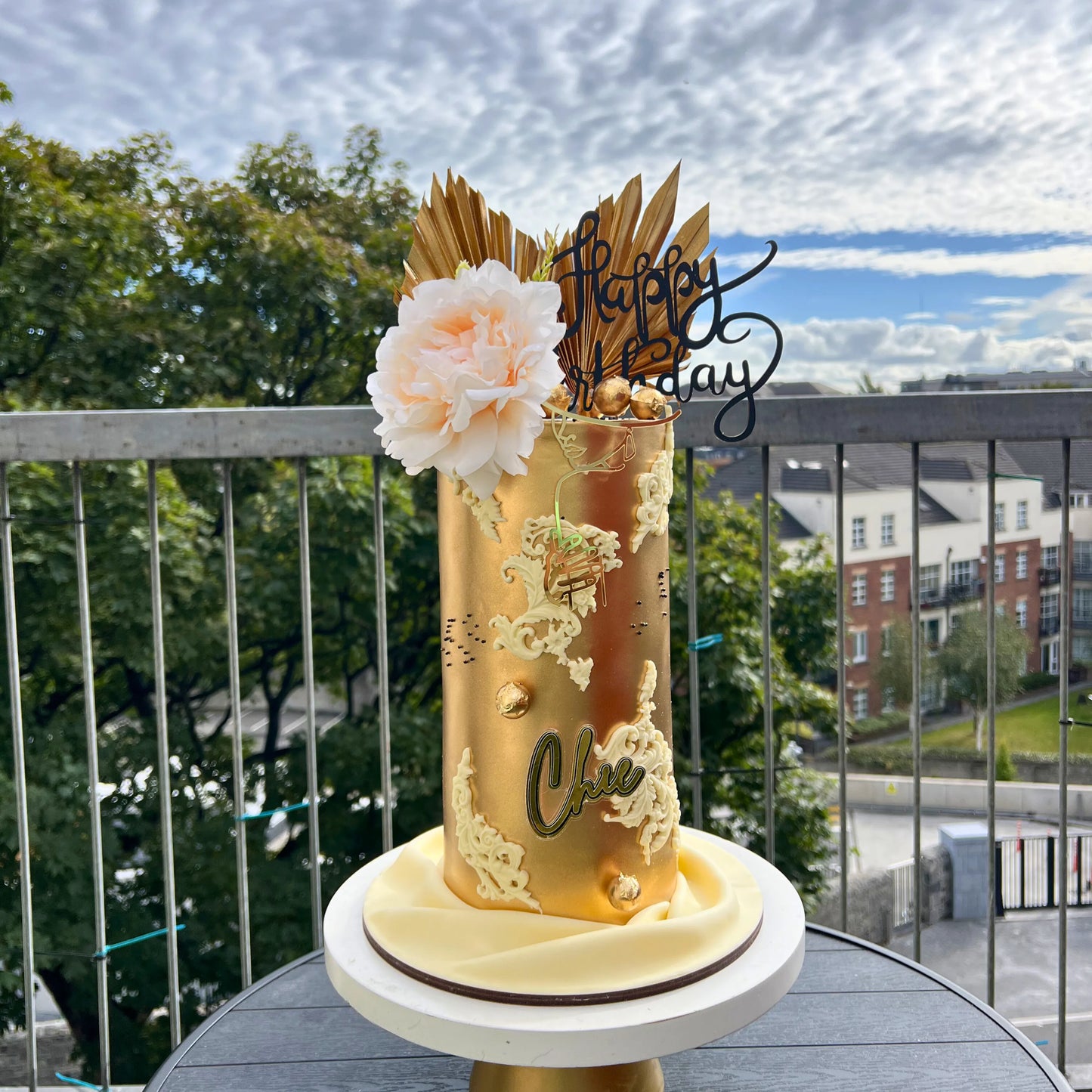 Gold birthday cake with floral decorations on a balcony with a view of trees and buildings.